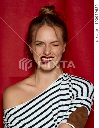 Smiling, positive vibe. Portrait of young beautiful girl with red lips and hair bun, in striped shirt posing against red studio background. Concept of beauty and fashion 100059689