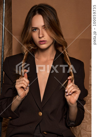 Tender serious natural look. Studio portrait of young beautiful girl in stylish dark suit posing against light brown background. Concept of beauty and fashion 100059735