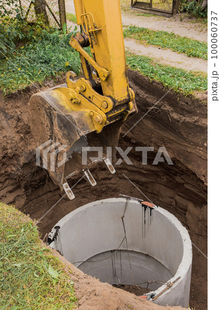 Excavator with a bucket, lowering into the pit on steel cables concrete sewer ring. Construction or repair of sewer home 100060737