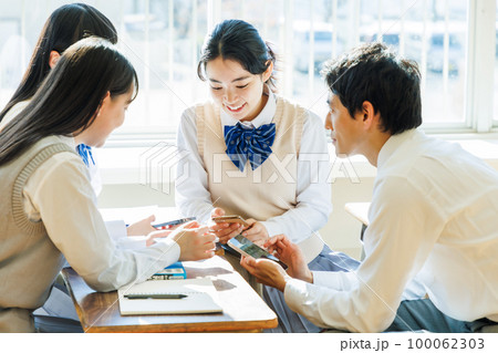 教室でクラスメイトとスマホを見ながら話す高校生 100062303