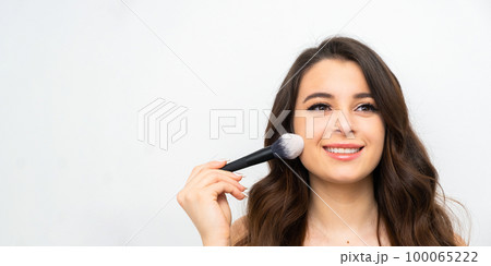 Banner with smiling young woman holding a cosmetic brush near her face. Makeup artist applying face powder. Banner with smiling young woman holding a cosmetic brush near her face. Makeup artist applying face powder. 100065222