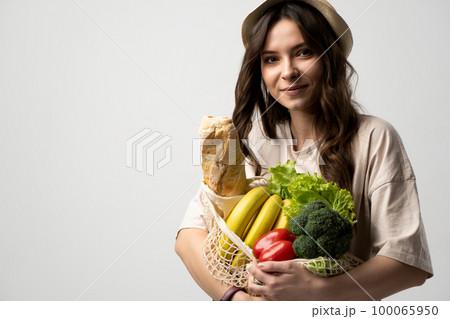 Eco friendly positive young woman in beige oversize t-shirt holding reusable mesh cotton eco bags for shopping with groceries on white background. 100065950