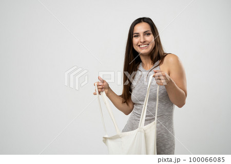 Cheerful millennial woman showing white eco bag to camera standing over white studio background. Lady holding cotton shopper handbag. fashion and ecology concept. 100066085