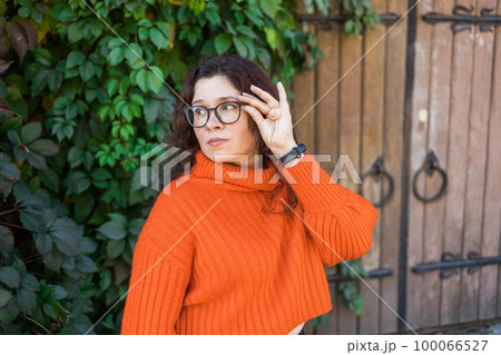 Portrait of carefree young woman smiling with big door background. Cheerful latin girl wearing eyeglasses in the city. Happy brunette woman with curly hair and spectacles smiling. Copy space and empty 100066527