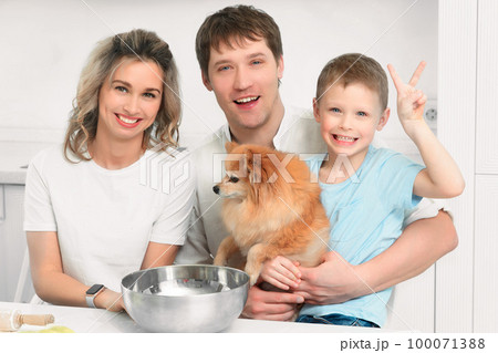 Cheerful happy family of three together with Spitz dog sit at kitchen table and laugh. Portrait of Cheerful happy family of three together with Spitz dog sit at kitchen table and laugh. Portrait of 100071388
