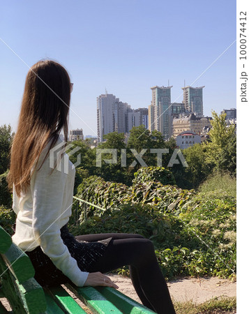 A beautiful brunette woman with long hair sits sideways and looks out over the city of Kiev in Ukraine in summer or spring. A beautiful brunette woman with long hair sits sideways and looks out over the city of Kiev in Ukraine in summer or spring. 100074412