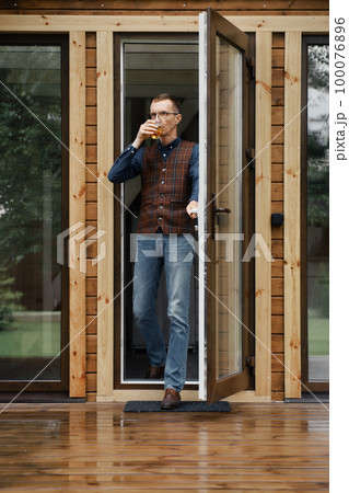 Middle age man comes out on the terrace of a small wooden house with a glass in hand Middle age man comes out on the terrace of a small wooden house with a glass in hand 100076896
