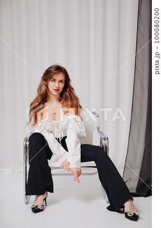 a beautiful woman with long hair in a white top and black pants on a chair. 100080200