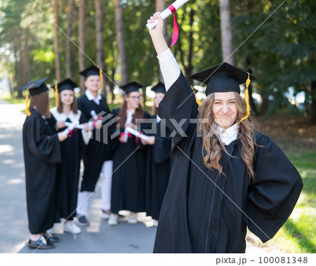 Group of happy students in graduation gowns outdoors. A young girl with a diploma in her hands in the foreground. 100081348