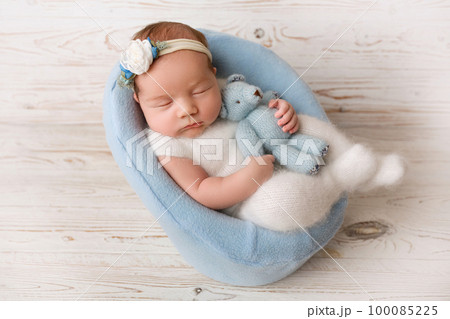 Top view of a newborn baby girl sleeping in a white jumpsuit with a blue teddy bear, in a blue felt chair. On a light wooden background. Beautiful portrait of a little girl 7 days, one week. Top view of a newborn baby girl sleeping in a white jumpsuit with a blue teddy bear, in a blue felt chair. On a light wooden background. Beautiful portrait of a little girl 7 days, one week. 100085225