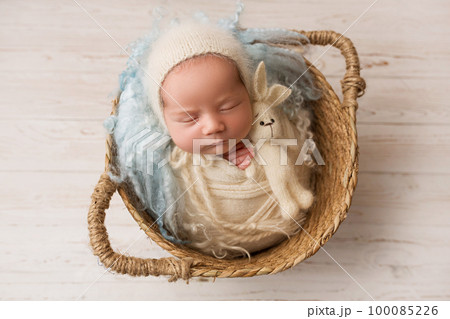 Top view of a newborn baby girl sleeping in a white cape with a blue bandage and a flower on her head, in wicker basket with blue fur and a rabbit. Beautiful portrait of a little girl 7 days, one week Top view of a newborn baby girl sleeping in a white cape with a blue bandage and a flower on her head, in wicker basket with blue fur and a rabbit. Beautiful portrait of a little girl 7 days, one week 100085226