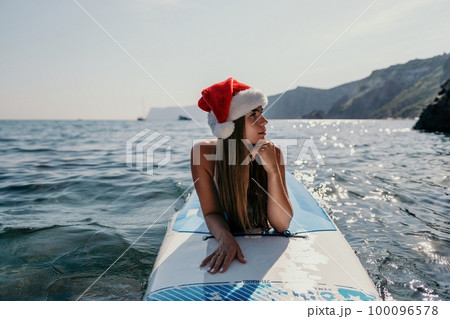 Woman sea sup. Close up portrait of happy young caucasian woman with long hair in Santa hat looking at camera and smiling. Cute woman portrait in a white bikini posing on sup board in the sea Woman sea sup. Close up portrait of happy young caucasian woman with long hair in Santa hat looking at camera and smiling. Cute woman portrait in a white bikini posing on sup board in the sea 100096578