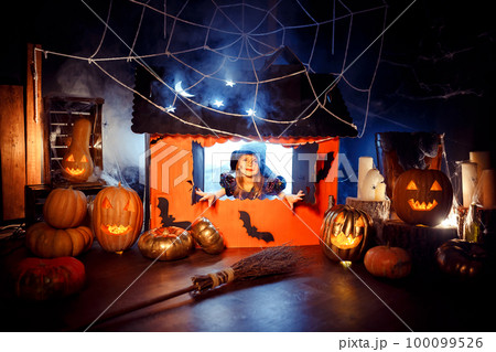 Little cute girl in witch costume looks out of the window of magic cardboard house. Halloween party  100099526