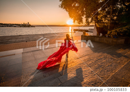 Sunrise red dress. A woman in a long red dress against the backdrop of sunrise, bright golden light of the sun's rays. The concept of femininity, harmony. 100105833