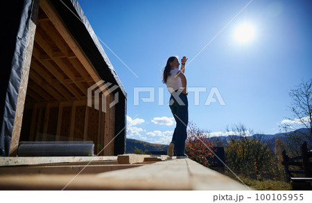 Happy woman on construction site standing on terrace at unfinished wooden frame house on sunny day. Happy woman on construction site standing on terrace at unfinished wooden frame house on sunny day. 100105955