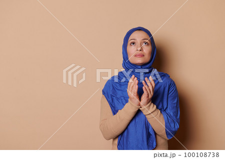 Peaceful Arab Muslim woman in blue hijab, looking up with gratitude, doing a day pray with folded hands, showing her believe in God isolated on beige backdrop. Islam. Religion. Middle-Eastern culture. 100108738