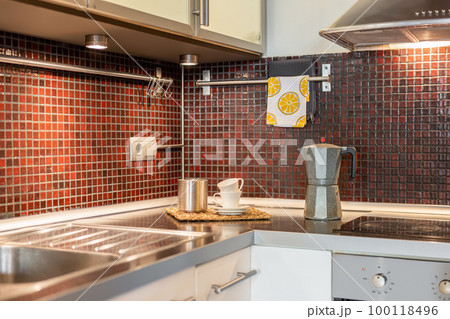 Fragment of kitchen island in corner and piece of sink with a stainless steel tray and coffee maker on the background of burgundy tiles and wall cabinets. Concept of convenient organization of space Fragment of kitchen island in corner and piece of sink with a stainless steel tray and coffee maker on the background of burgundy tiles and wall cabinets. Concept of convenient organization of space 100118496