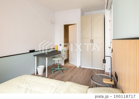 Compact but cozy bedroom with a double bed, a table, a chair and a lamp in a bright interior with a built-in wardrobe and an open door to the bathroom. Hotel room concept 100118717