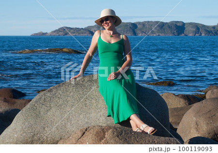 Woman tourist in a straw hat and a green dress on the seashore on a bright sunny day 100119093