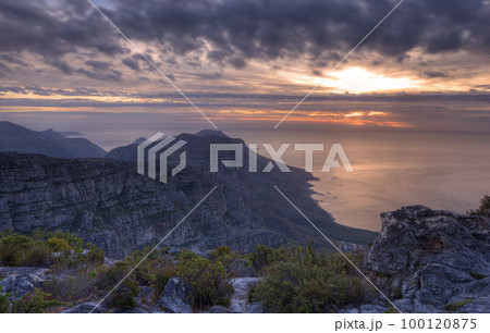 Above view of a mountain coastline at sunset in South Africa. Scenic landscape of dark clouds over a calm and peaceful ocean near Cape Town with the sun behind grey clouds in the sky and copy space Above view of a mountain coastline at sunset in South Africa. Scenic landscape of dark clouds over a calm and peaceful ocean near Cape Town with the sun behind grey clouds in the sky and copy space 100120875