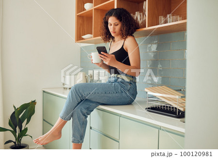 Afro woman on phone typing, texting or reading online fake news at home drinking coffee or tea. Trendy, young and stylish female sitting on kitchen counter browsing the internet and social media. 100120981