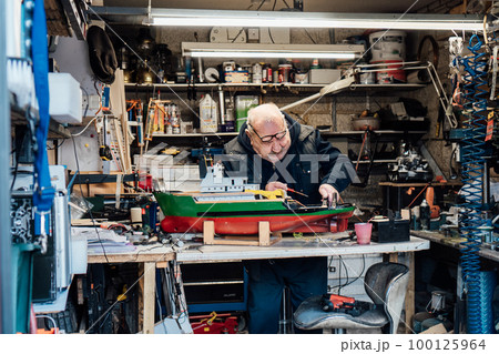 Portrait of a senior man, pensioner holding made handicraft ship model with background of his garage with different materials. Retired elderly man hobby. Active Retirement lifestyle. Selective focus. Portrait of a senior man, pensioner holding made handicraft ship model with background of his garage with different materials. Retired elderly man hobby. Active Retirement lifestyle. Selective focus. 100125964