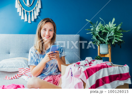 Smiling woman using smartphone to find places where to donate or sale clothes. Decluttering, Sorting clothes and Cleaning Up. Conscious consumer, sustainability. Reuse, second hand, Charity concept 100126015