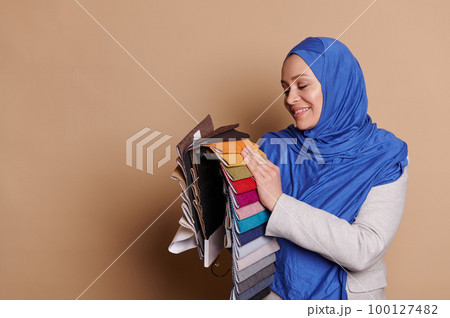 Smiling Muslim woman in blue hijab, sales manager, interior designer or client in the upholstered furniture store, putting her hand on the upholstery textile color swatches, isolated beige background 100127482