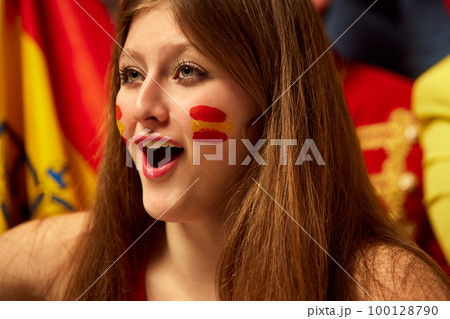 Close-up image of girl, football, soccer fans emotionally watching match, cheering up favourite team. Fans wearing colorful scarves and flags Close-up image of girl, football, soccer fans emotionally watching match, cheering up favourite team. Fans wearing colorful scarves and flags 100128790