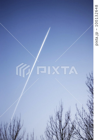 Jet airplane contrail above leafless trees against a clear blue sky background with copyspace. View of a distant passenger plane flying at high altitude leaving a long white smoke trail behind 100132648