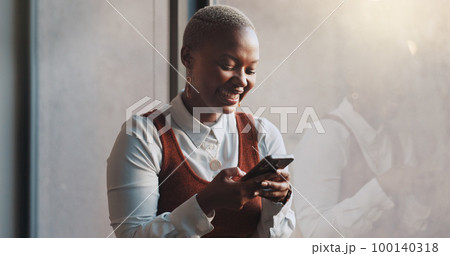 Happy, smile and face of a black woman on a phone while standing by the window in office. Technology, happiness and African female employee networking on social media or the internet on break at work 100140318