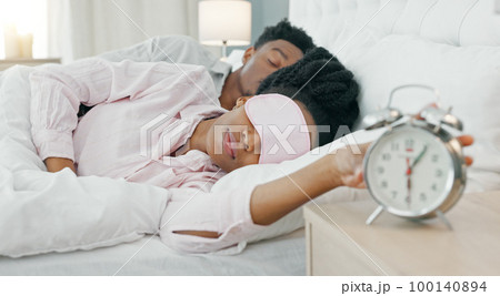 Black woman waking up in bed to switch off her alarm clock with a sleep mask in her house. Lazy, sleepy and African girl sleeping in her comfortable bedroom with an early morning get up time at home. 100140894