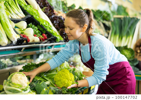 Middle-aged saleswoman putting cauliflower on food stall in greengrocery 100147702