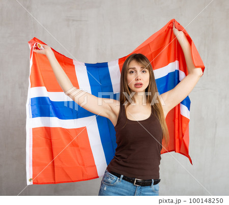 Upset patriotic young female student holding state flag of Norway against gray wall background indoors 100148250
