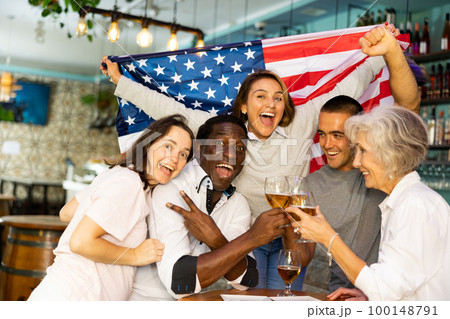Happy sport fans holding flag of United States of America, celebrating and drinking alcoholic drinks in pub, man showing victory sign Happy sport fans holding flag of United States of America, celebrating and drinking alcoholic drinks in pub, man showing victory sign 100148791