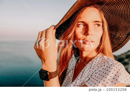 Portrait of happy young woman wearing summer black hat with large brim at beach on sunset. Closeup face of attractive girl with black straw hat. Happy young woman smiling and looking at camera at sea. Portrait of happy young woman wearing summer black hat with large brim at beach on sunset. Closeup face of attractive girl with black straw hat. Happy young woman smiling and looking at camera at sea. 100153206