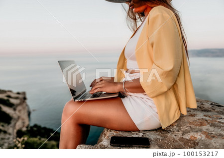 Digital nomad, Business woman working on laptop by the sea. Pretty lady typing on computer by the sea at sunset, makes a business transaction online from a distance. Freelance, remote work on vacation 100153217