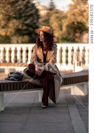 Outdoor fashion portrait of young elegant fashionable brunette woman, model in stylish hat, choker and light raincoat posing at sunset in European city. 100162376