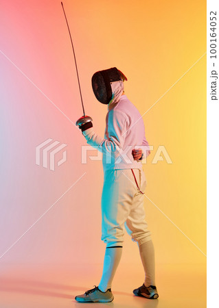 Young man, male fencer with sword practicing in fencing over gradient pink-yellow background in neon light. Sportsman shows fencing technique 100164052