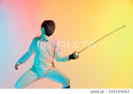 Young man, male fencer with sword practicing in fencing over gradient pink-yellow background in neon light. Sportsman shows fencing technique 100164062