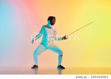 Young man, male fencer with sword practicing in fencing over gradient pink-yellow background in neon light. Sportsman shows fencing technique 100164063