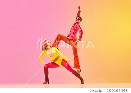 Young stylish emotional man and woman, professional dancers in retro style clothes dancing disco dance over pink-yellow background. 1970s, 1980s fashion, music concept 100165448