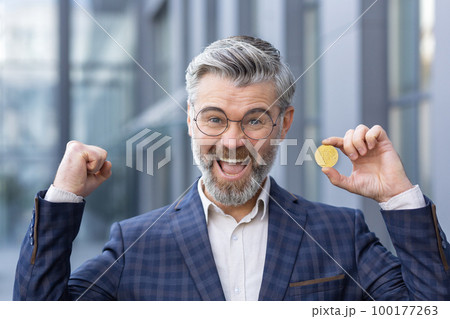 Close-up photo. A senior man, businessman standing outside office and holding bitcoin, gold coin, cryptocurrency in hand. He looks at the camera. shows a winning gesture with his hands, smiles. 100177263