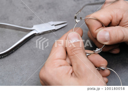 Closeup of optician's hands repairing broken eyeglasses frame using special pliers in clinic office Closeup of optician's hands repairing broken eyeglasses frame using special pliers in clinic office 100180058