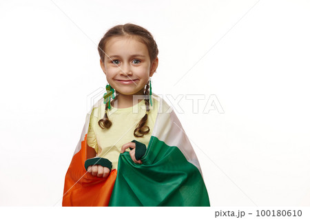 Mischievous lovely little girl with two funny pigtails, wearing earring in the shape of clover leaves, posing wrapped in Ireland flag on isolated white background with copy space. Saint Patrick's Day 100180610