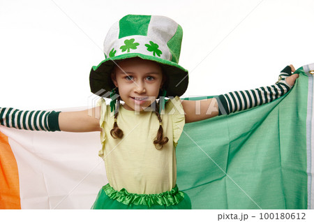 Adorable Caucasian little child girl dressed as Leprechaun in hat with clover leaves and striped gloves, smiling looking at camera, carrying Ireland flag, white background. Saint Patrick's day concept 100180612