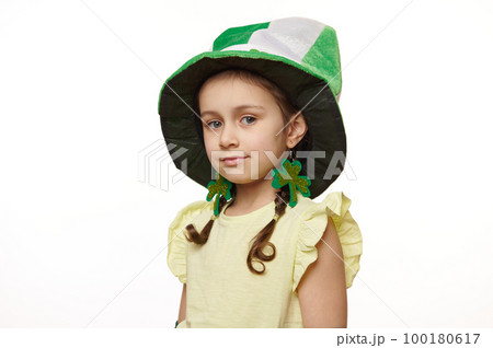 Studio shot of stylish Irish little baby girl dressed like a Leprechaun for St Patrick's Day party, wearing clover leaves earrings and green hat, smiles looking at camera, isolated on white background 100180617
