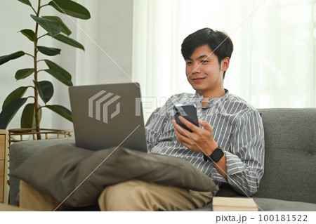 Cheerful asian man hand holding mobile phone and looking at laptop screen. People, technology and lifestyle concept 100181522