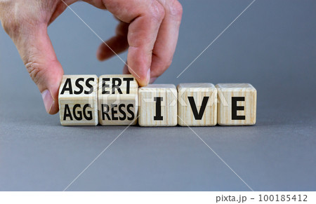 Aggressive or assertive symbol. Businessman turns wooden cubes, changes the word Aggressive to Assertive. Beautiful grey background, copy space. Business, psychological aggressive assertive concept. Aggressive or assertive symbol. Businessman turns wooden cubes, changes the word Aggressive to Assertive. Beautiful grey background, copy space. Business, psychological aggressive assertive concept. 100185412