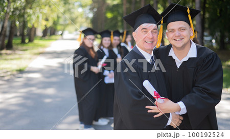 A group of graduates in robes outdoors. An elderly man and a young guy congratulate each other on receiving a diploma. 100192524